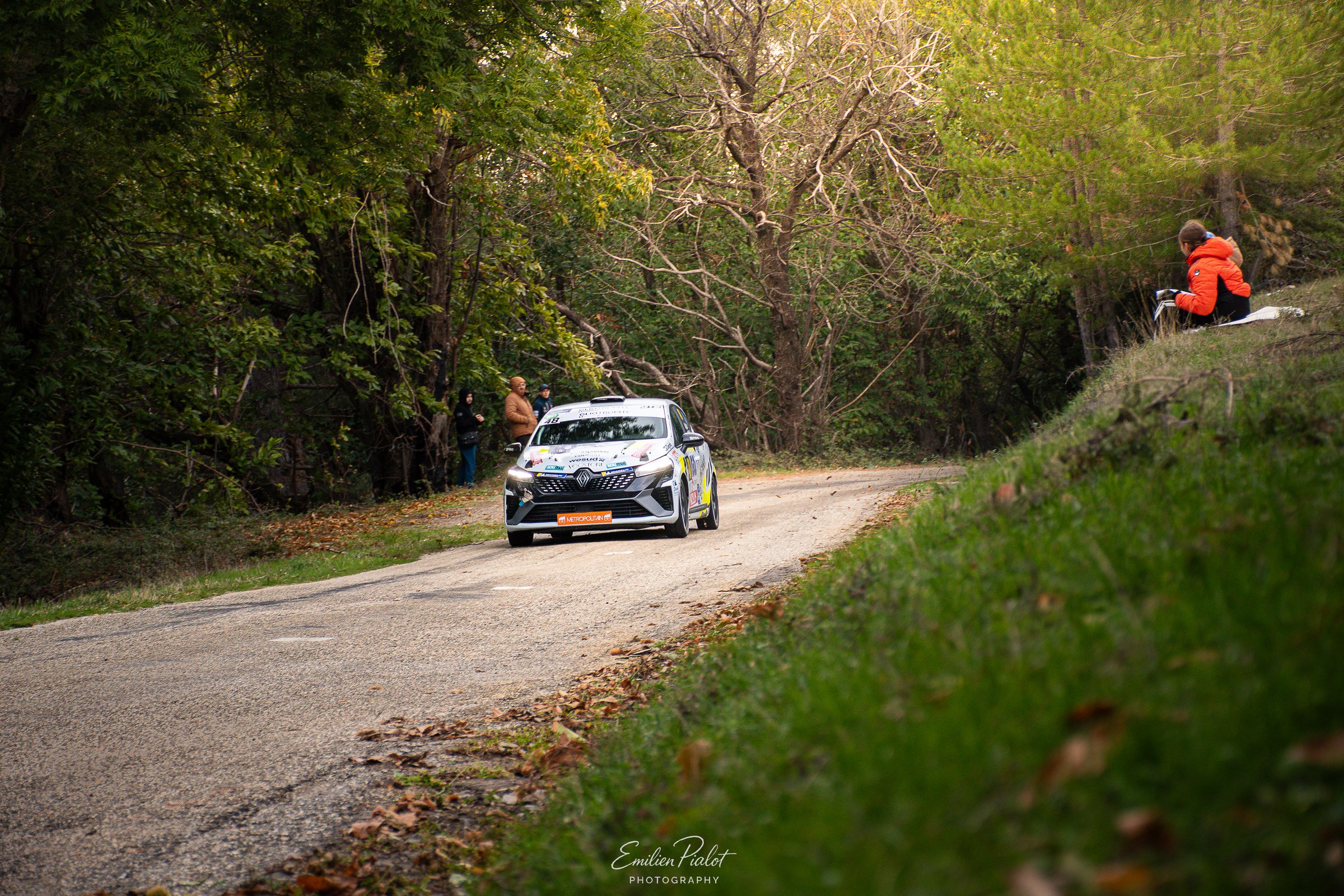 Voiture de rallye bleue et jaune phares allumés sur un chemin de terre sous les arbres