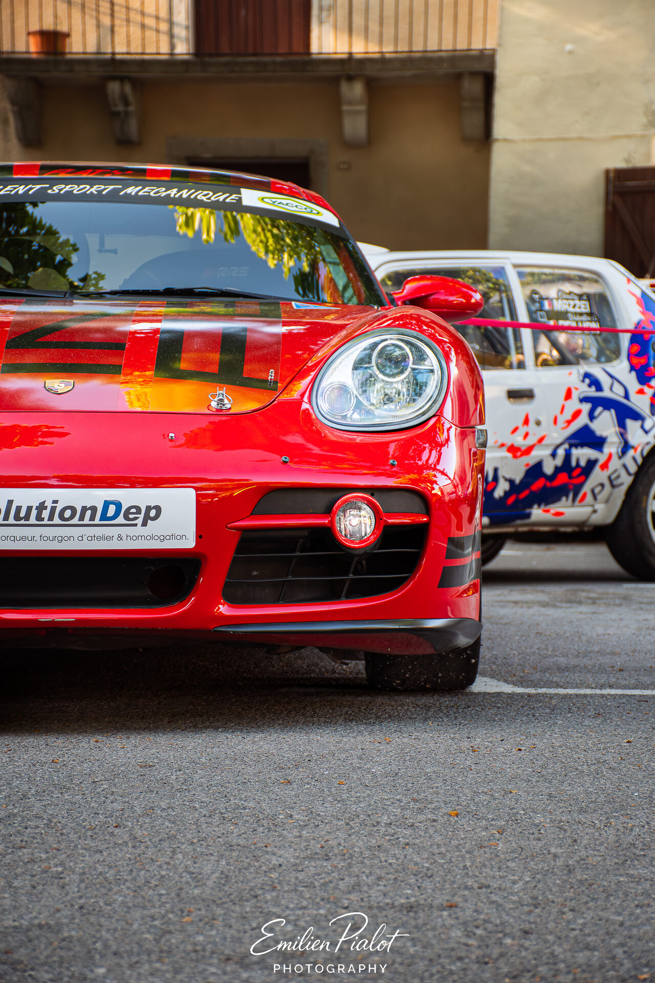 Porsche Cayman rouge devant une Peugeot 205 blanche et bleue, ruelle du village