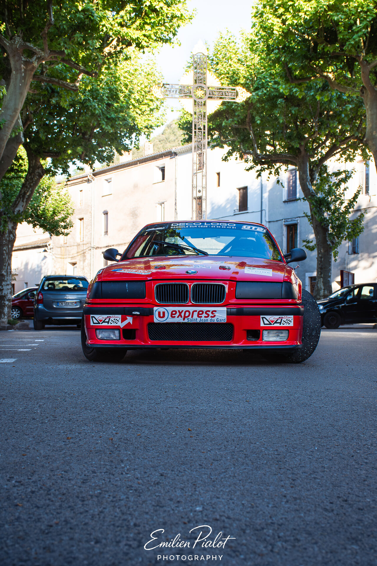 BMW M3 E36 rouge vue de face, croix en fer forgé sous les platanes